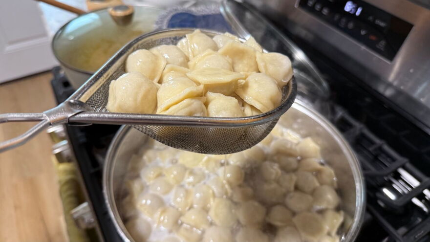 A pot full of dumplings is cooking. Above the pot, someone is holding a strainer full of dumplings.