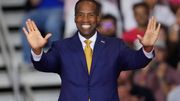 A smiling man in a suit extends his arms out in front of him during a rally.