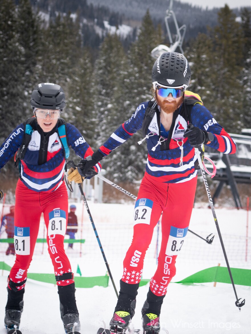 Two people in ski mountaineering gear race up a hill on a snowy racecourse.
