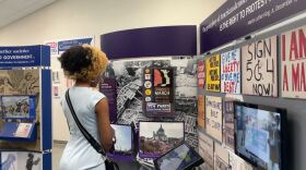 A woman's back is turned to the camera as she looks at various protest materials, slogan signage and museum plaques. She is wearing a blue dress and there is a TV on the right wall playing a protest video.