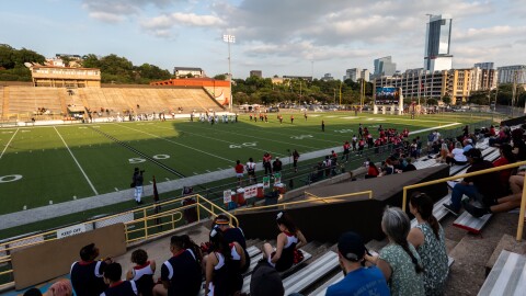 A high school football stadium with fans in stands and a portion of the city skyline in the background.