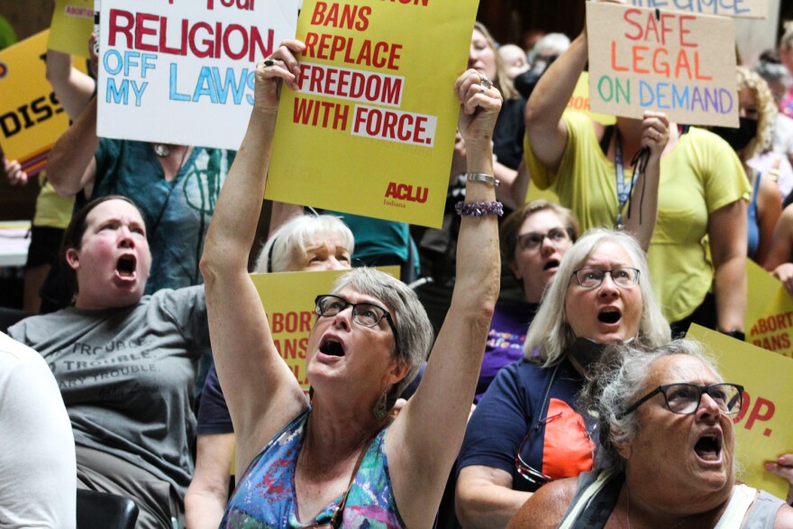 A group of people rally at the Indiana Statehouse in support of abortion rights. They're holding signs, which include the words "Abortion bans replace freedom with force" and "Safe, legal, on demand." The group of people are all looking up and shouting.