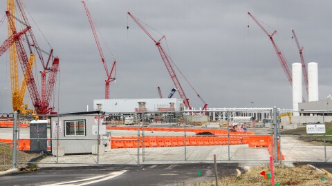 Construction cranes rise above a construction site blocked off by a large chain-link gate. A sign reads, "gate closed." 