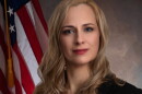 A woman with long blonde hair is wearing a black blazer over a red blouse and posing next to an American flag.