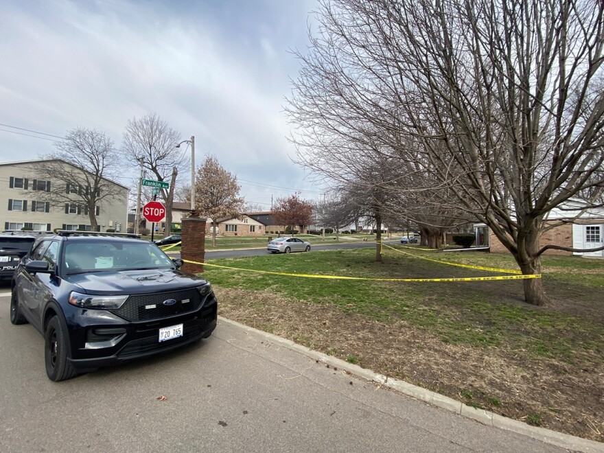 Crime scene tape along several homes and apartment buildings in an urban neighborhood with a police car parked at the curb