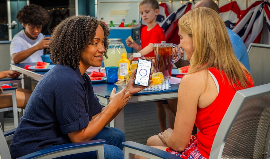 Two women sit at an outdoor table during a summer cookout, looking at an energy-tracking app on a smartphone. In the background, children and an adult eat watermelon and corn at a table decorated with patriotic bunting, lemonade, and iced tea.