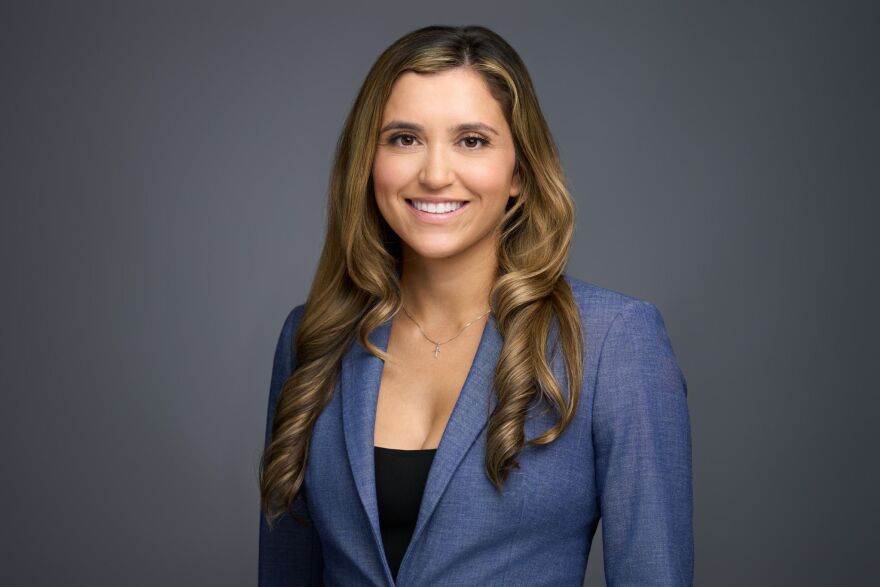 Woman with long brown hair standing in front of a grey background wearing a black top and blue blazer
