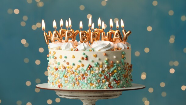 Beautiful birthday cake with burning candles on stand against festive lights
