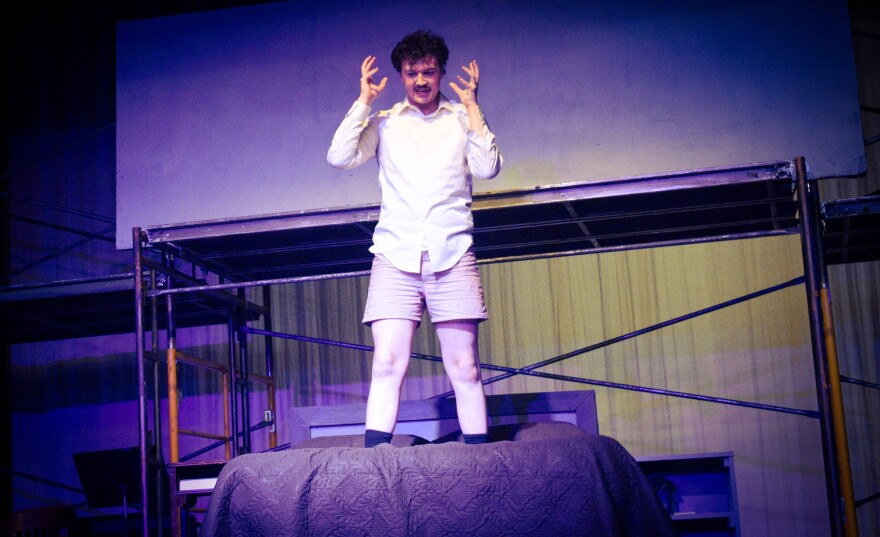 A person in a white shirt and boxers stands on a bed, raising their hands and making an intense expression, with stage scaffolding and dramatic lighting in the background.