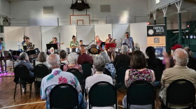 A steel band performs inside a community hall while an audience watches from rows of chairs.