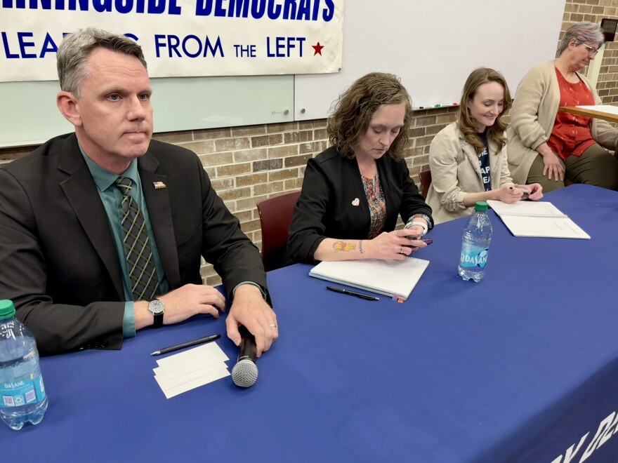 A man and two women sit at a table with a blue tablecloth. The man on the left is holding a microphone.