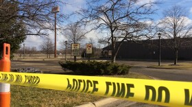 Police tape surrounds a black building with an empty parking lot. The grass is short and the trees are bare, denoting the winter season despite the lack of snow. The sign for Cracker Barrel can be seen at the top right of the image, partially covered by the branches of a tree in the foreground. 