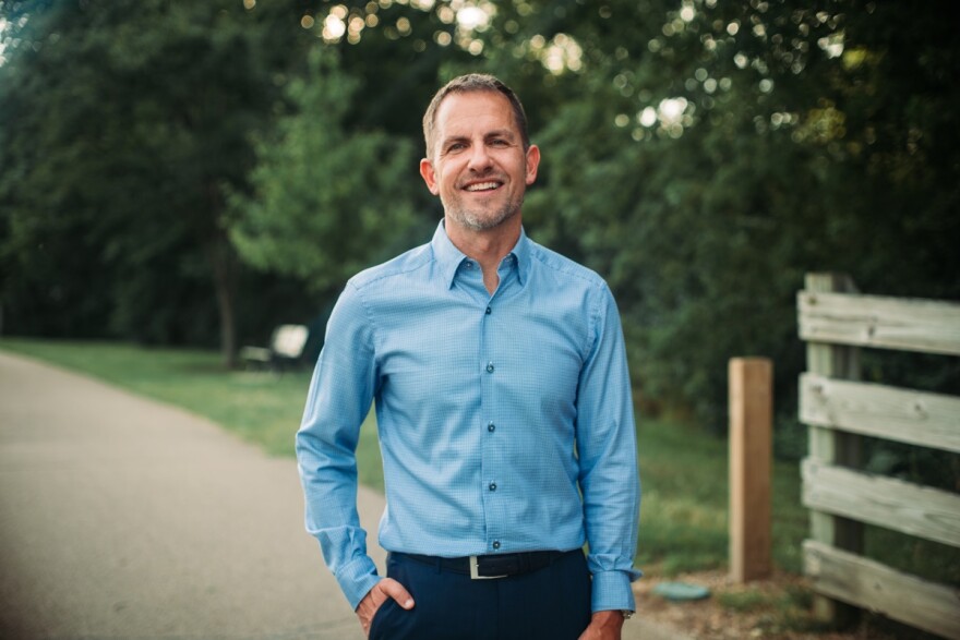 Man in light blue long-sleeve buttoned-down shirt standing along a paved bike trail with trees in the background and a wooden fence to his left