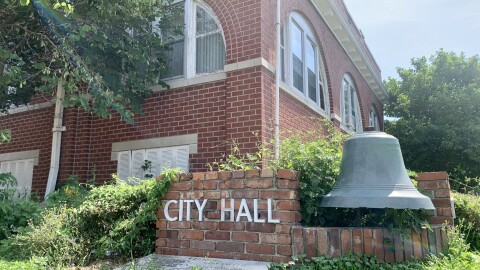 A close up of the city hall sign outside of Mexico Missouri. There is white lettering that spells "City Hall" on a brick background. There is also a large metal bell next to the sign. The red brick building of city hall is in the background. 
