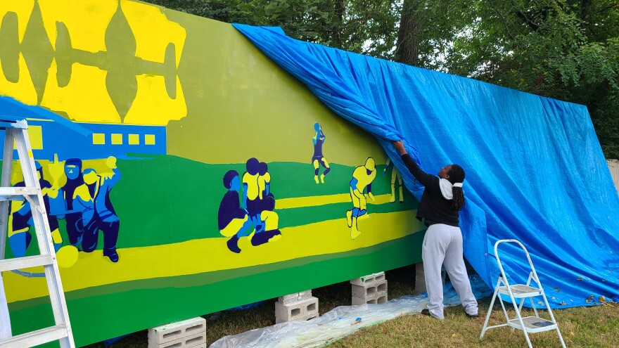 Artist Myntia Daniels rolls back the mural's protective tarp to resume painting after Thursday morning's rain showers.
