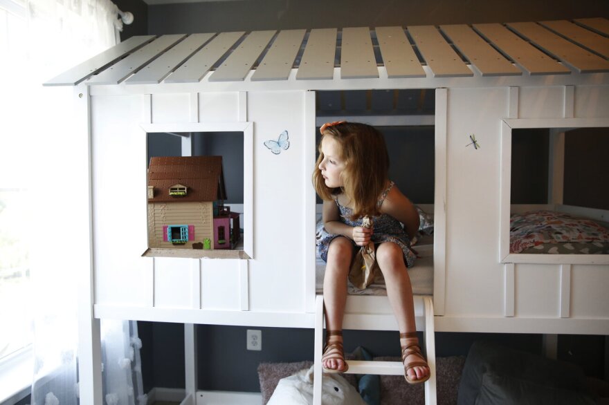 Maddie, 6, sits atop her lofted bed, which is styled like a tree house. She wears a patterned dress and brown sandals and looks toward a window.
