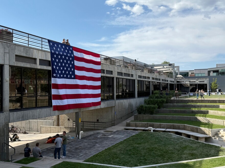 People gaze down at the site where Charlie Kirk was shot, above which hangs an American flag, at Utah Valley University, Monday, Sept. 15, 2025, in Orem, Utah.