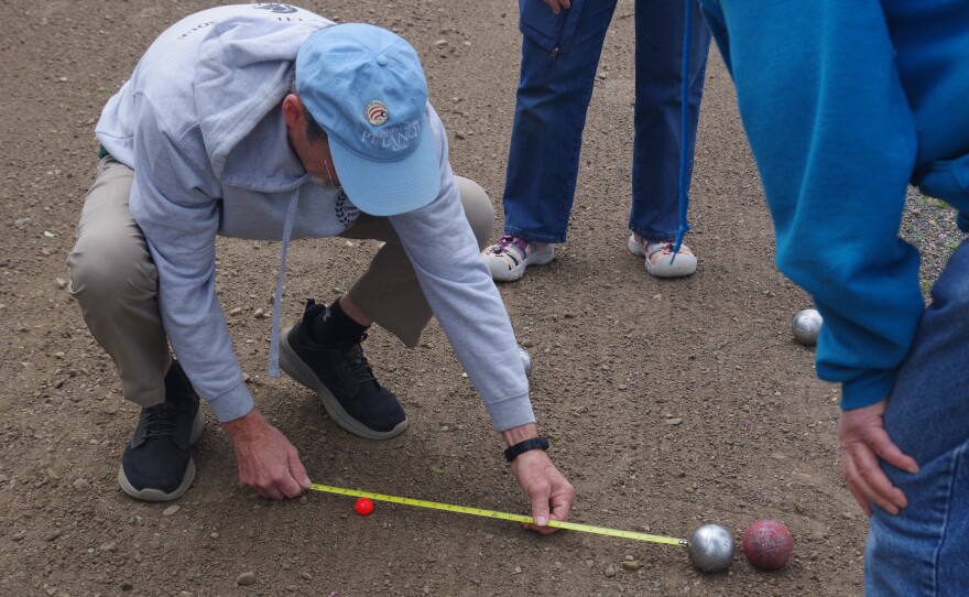 man crouches with measuring tape to measure distance between target and boules on a gravel surface, while several people look on