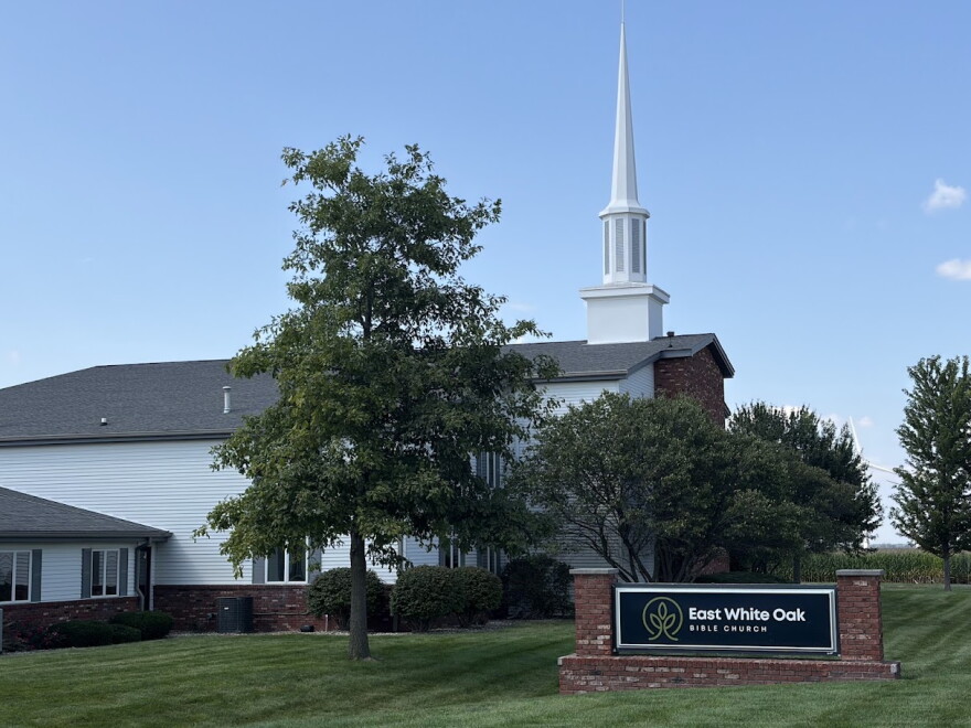 a sign reading "East White Oak Bible Church" is in the foreground, a white church steeple stands in the background