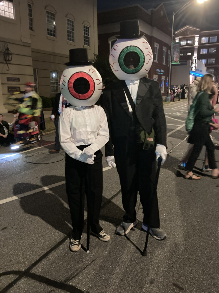A child and an adult, dressed in tuxedos on top hats an canes, with giant eyeballs as heads, stand in the street at an annual Halloween Parade.