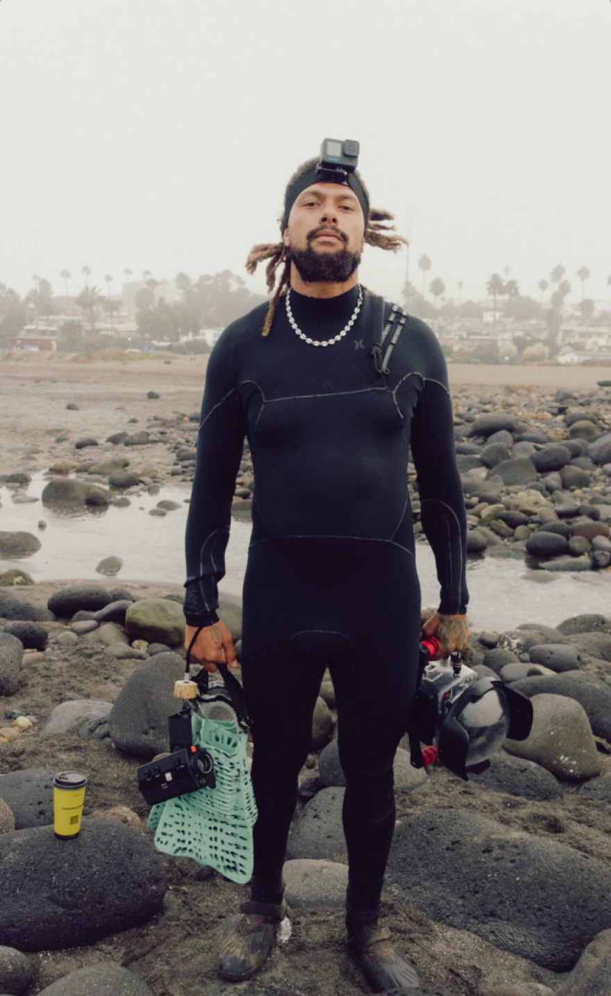 Kory Lamberts stands on rocky beach in a wetsuit holding dive equipment and cameras.
