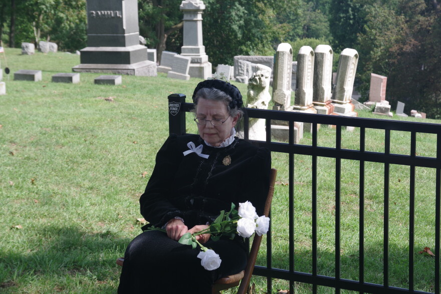 A woman in an early 20th century long black funeral gown and matching black bonnet sits on a chair with her eyes closed. She's holding a bouquet of white flowers, with a number of gravestones sitting behind her. 
