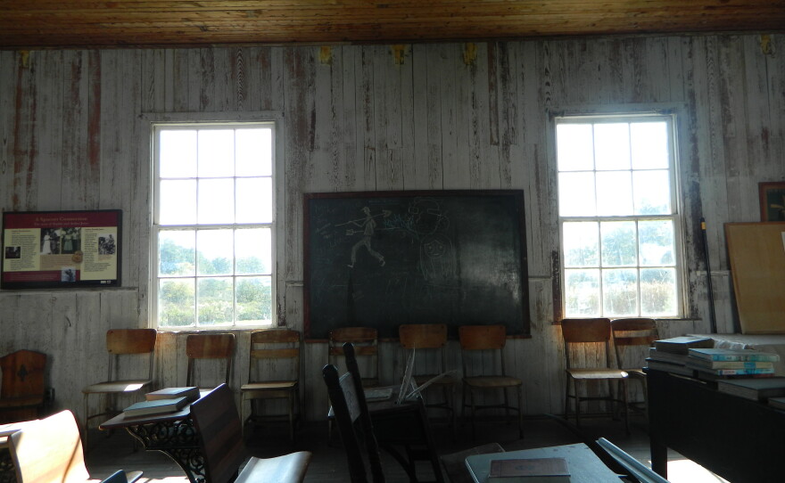 The Castle Haven schoolhouses. Old school desks in the foreground, chairs lined up against the wall where a blackboard hangs with doodles scribbled onto it.