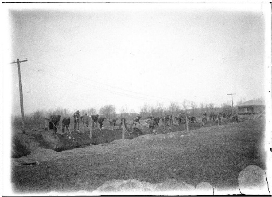 About twenty Thomas Indian School students dig a large trench with shovels along a field in a black and white photo taken sometime between 1900 and 1930.