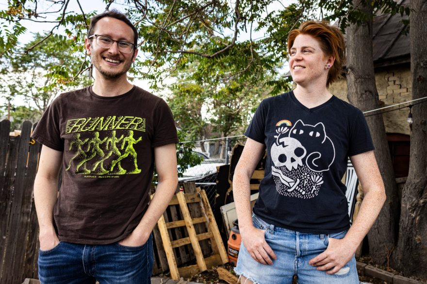 Two people stand outside in a backyard. One smiles at the camera while the other smiles at the first person. There is a worn-down fence and some wood supplies visible behind them both.