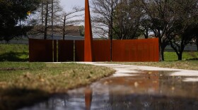 A burnt orange structure shaped like a sundial is seen from a distance in a Dallas park.