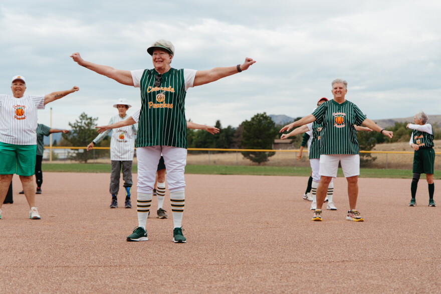 Senior women stand out in a baseball field a few feet apart from each other with their arms out in a T-pose, circling them. They are wearing green and white baseball jerseys that say "Colorado Peaches" on them.