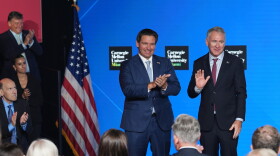Two men wearing suits wave as they stand on a stage next to an American flag, flanked by people clapping. 
