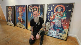 Man with tattoos all over his arms and a long grey beard squats in front of his artwork 