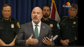 Un hombre calvo con traje habla desde un podio, flanqueado por agentes de la ley.