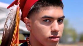 Coy-a-dee (Water Boy) Salomon poses for a photo in a red graduation robe and a red graduation cap adorned with a brown and white bird's feather.