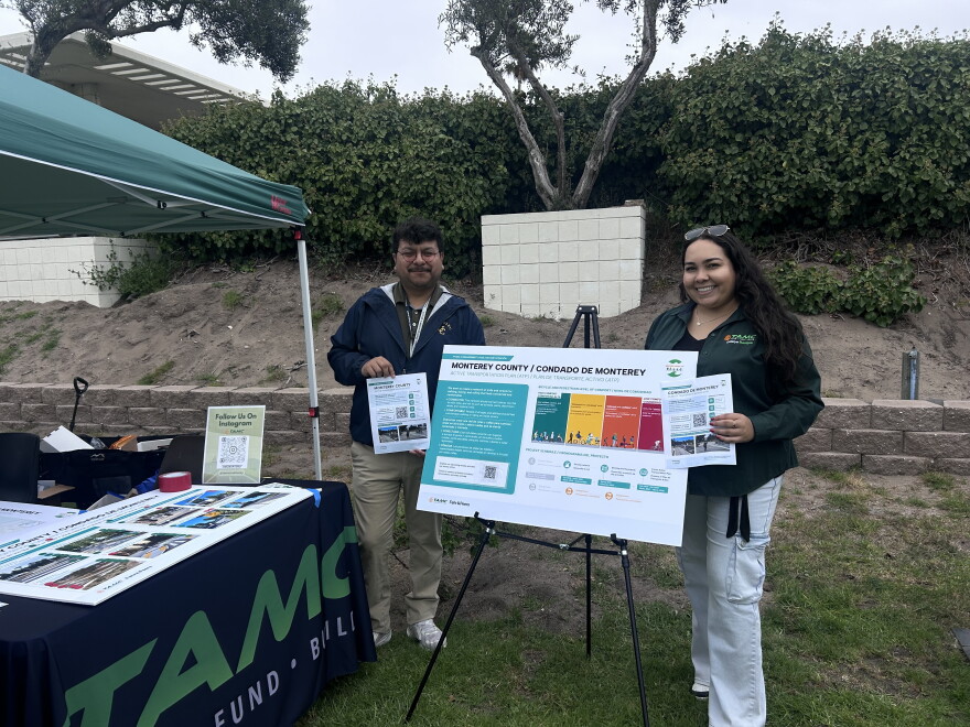 Two people standing holding flyers with a poster board on a stand between them. They are standing outside, a booth with a pop-up tent is to their left.