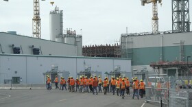 Wokers with orange vests and yellow hard hats make their way to a large grey nuclear waste facility.