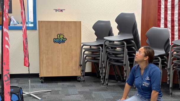 A young woman, sitting on the floor next to an open laptop and drone controller, watches a small drone float beneath a pink arch.