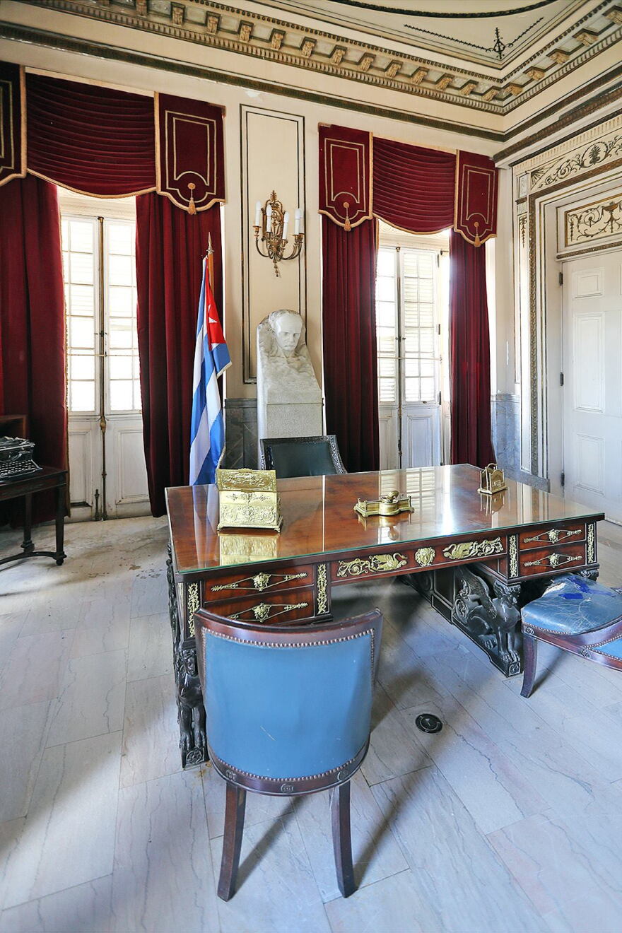 Two windows with heavy ornate drapery, the Cuban flag and a desk and two chairs, the former office of Cuban president Batista.