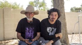 An elderly father and his adult son sit next to a tree in a backyard surrounded by a block wall. 