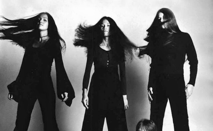 The Four women that make up the band Fanny, dressed in black. Three are standing, whipping their hair to the left, the fourth reclines on the floor.