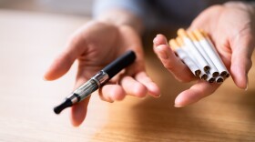 Woman's Hand Holding Vape And Tobacco Cigarettes Over Wooden Desk