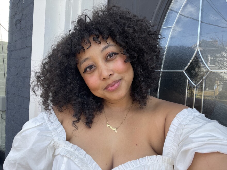 Minda Honey, a writer from Louisville, smiles into the camera. Honey, a Black woman, wears a white blouse, gold necklace and curly hair.