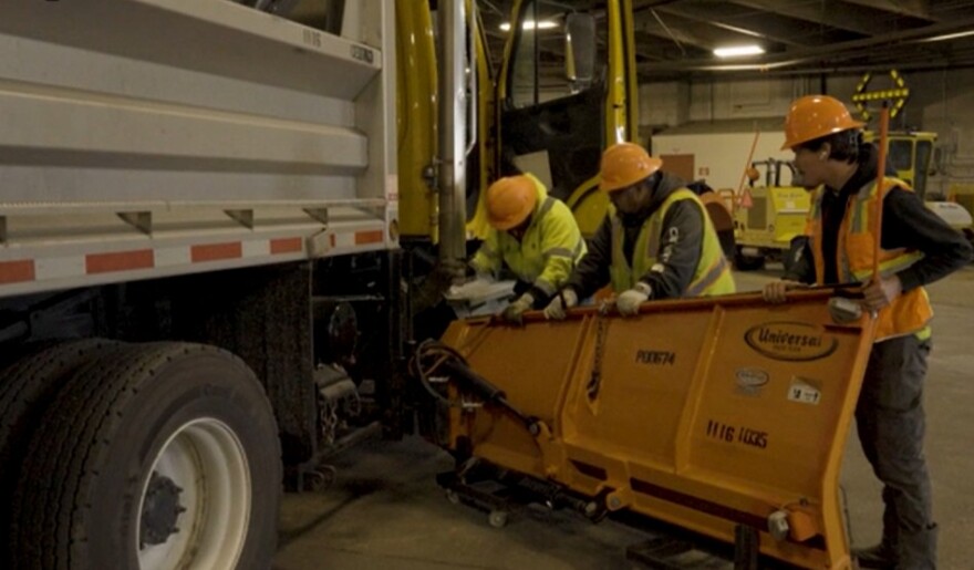 Three men in hard hats and hi-viz jackets hold a plow while one hammers at its attachment to a big truck.