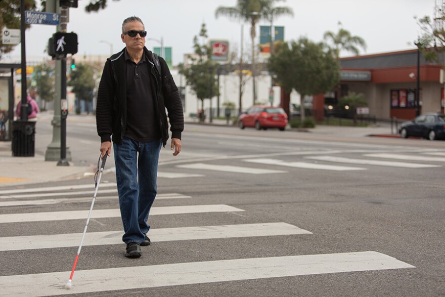 A man crosses a street with the aid of a white cane.