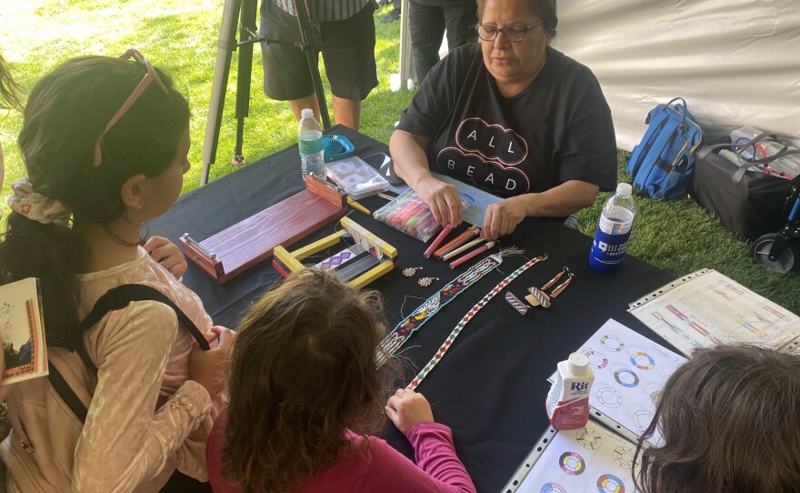 Three children are standing in front of a table hosted by the craft store All Bead Up to listen to how a bead worker creates her crafts.