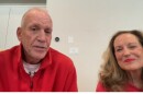 Doug and Kathy Collins seated in an office area with a white wall and cabinets behind them and both are wearing red sweaters