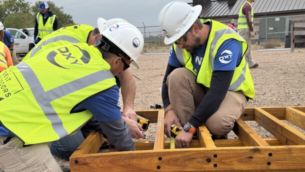Three construction workers in yellow vests are building a wooden structure on the ground.