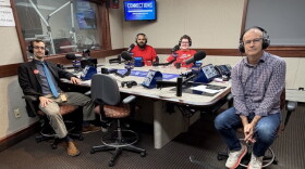 Four smiling people wearing headphones sit at a table in a radio talk studio: a man front left has short brown hair and a brown mustache and beard and is wearing glasses, a dark blue button-down shirt and lighter blue tie under a black suit coat, khakis, dark socks, and brown shoes; a man back left has short black hair and a black mustache and beard and is wearing a red polo shirt; a woman back center has short brown hair and is wearing glasses and a red graphic t-shirt; a man at right has very short dark hair and is wearing glasses, a purple and white checked button-down shirt, jeans, and white and red sneakers