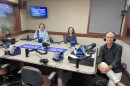 Three people wearing headphones sit at a table in a radio talk studio: a man at left has short grey hair and is wearing a blue plaid button-down shirt; a woman at center has long brown hair and is wearing eyeglasses, a blue blazer and blue blouse; a man at right has very short grey hair and is wearing eyeglasses, a black button-down shirt, white pants and sneakers.